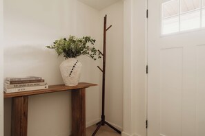 Una stanza d'angolo con pavimento in legno, una porta bianca, un appendiabiti, un vaso bianco con piante verdi e una pila di libri su un tavolo in legno.