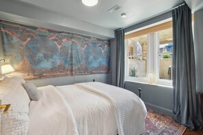 Une chambre avec une grande tapisserie représentant une carte du monde, un lit avec une literie blanche, une fenêtre avec des rideaux et une vue sur l’extérieur.
