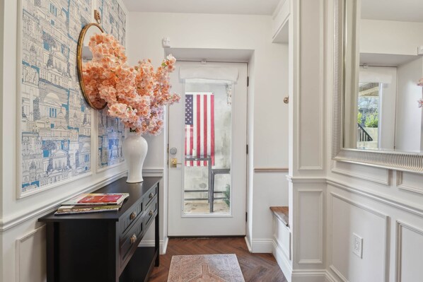 Un couloir avec une porte, un miroir, un vase de fleurs, un tapis et un mur orné d’une reproduction d’un paysage urbain.