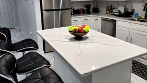 A modern kitchen with white cabinets, a stainless steel refrigerator, a marble countertop, and a bowl of fruit.