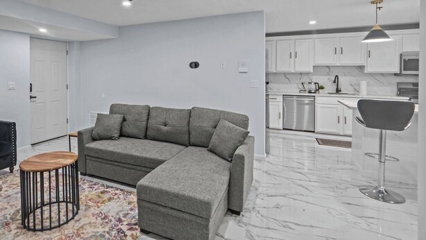 A modern living room with a grey sectional sofa, a small wooden table, and a floor lamp. The room has white walls, a marble floor, and a kitchen area with white cabinets and stainless steel appliances.