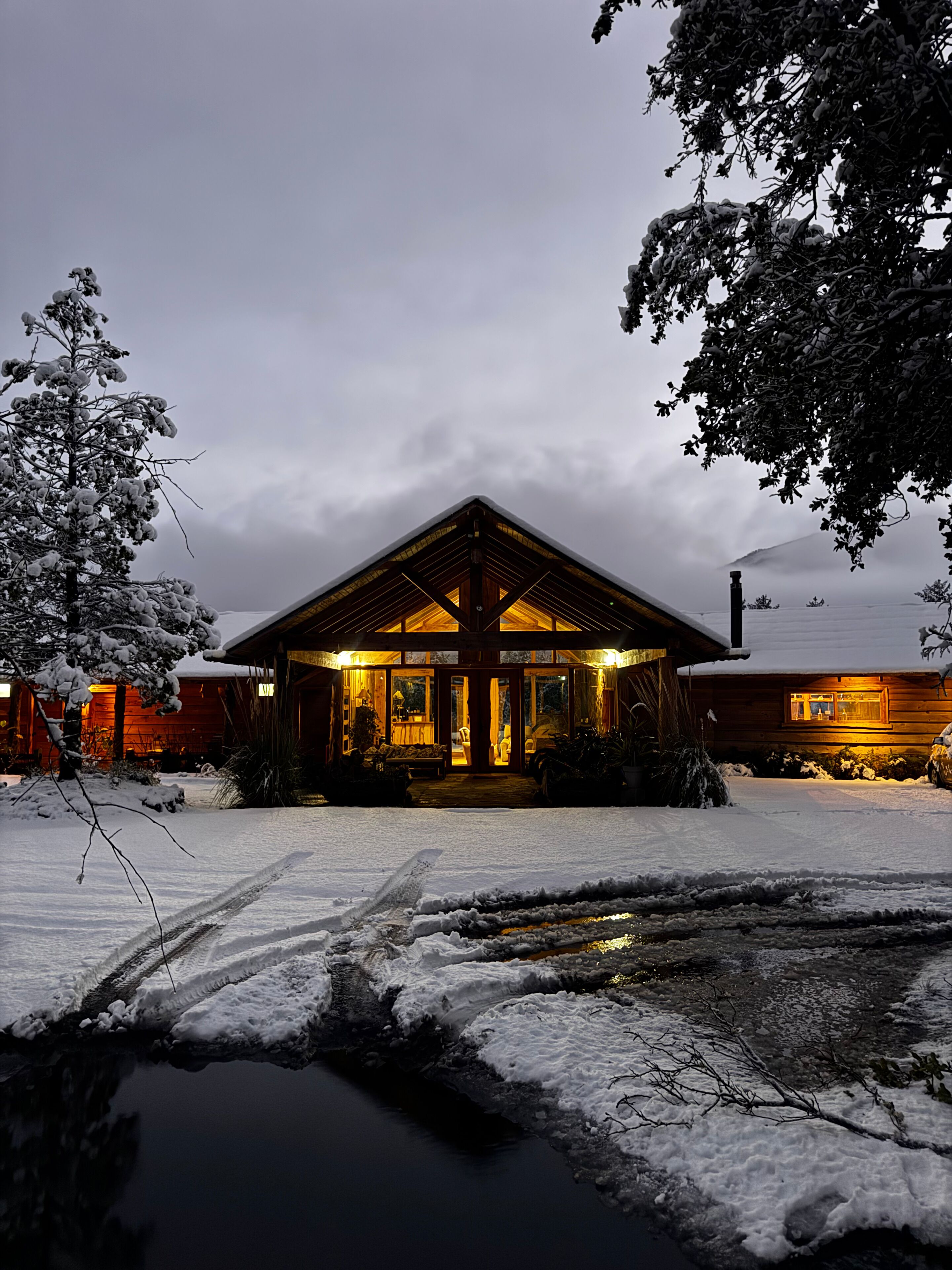 Una cabaña de madera con un porche cubierto, rodeada de terreno y árboles nevados.