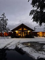 Una cabaña de madera con un porche cubierto, rodeada de terreno y árboles nevados.