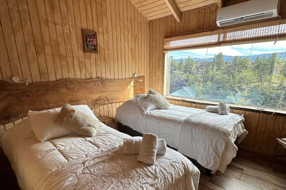 Habitación de cabaña de madera con dos camas, una ventana que da a árboles y un ventilador de techo.