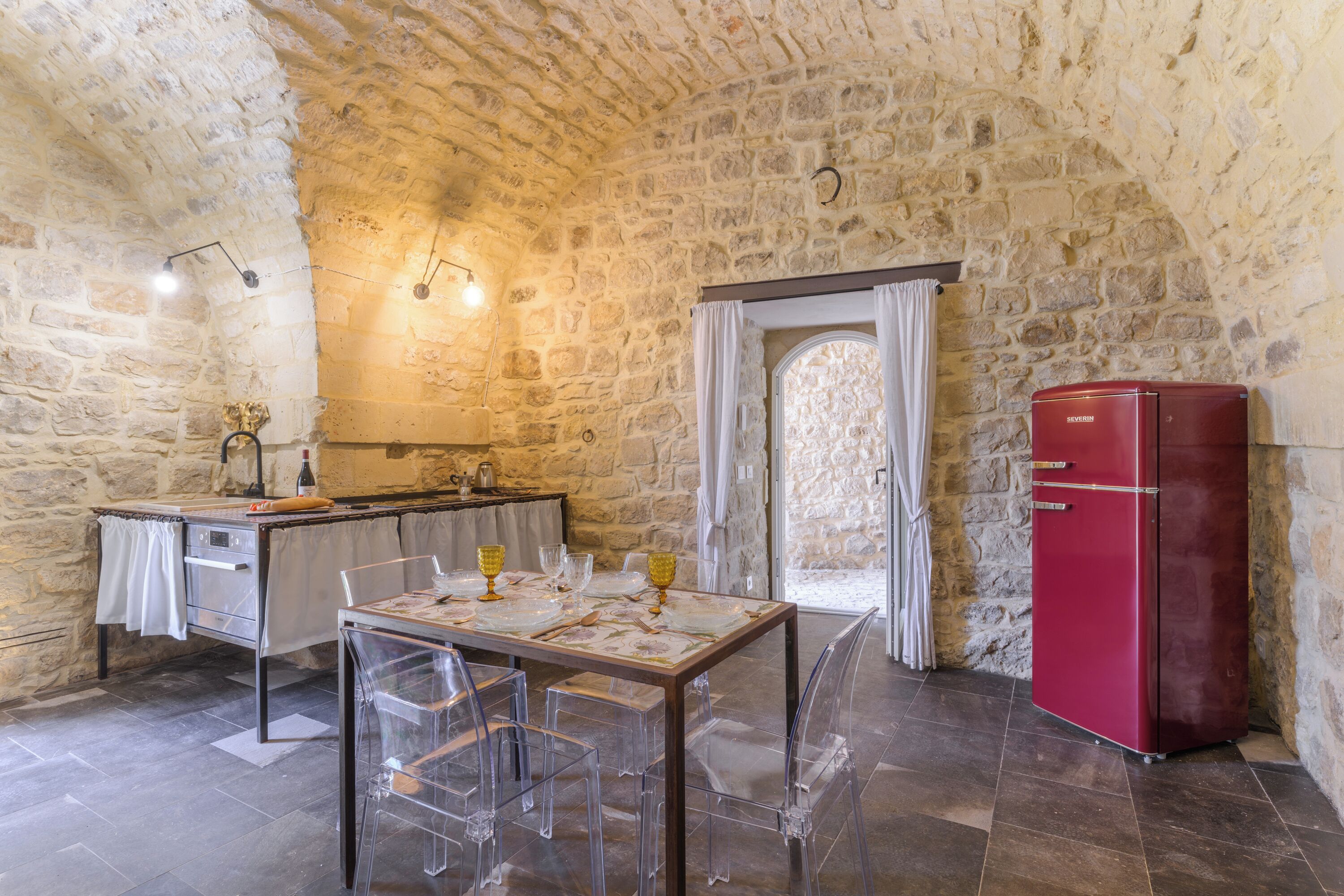 A stone-walled kitchen with a dining table set for two, a red refrigerator, and a window with curtains.