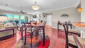 A spacious living room with a large window, a dining area with a wooden table and chairs, and a kitchen with a countertop and cabinets. - Bridgewater Pointe (Westlake Corner)