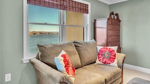 A living room with a beige sofa, two decorative pillows, a wooden cabinet, and a large window with a view of a body of water. - Bridgewater Pointe (Westlake Corner)