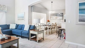 A modern living room with a blue sofa, a dining area with a table and chairs, and a kitchen area with bar stools.