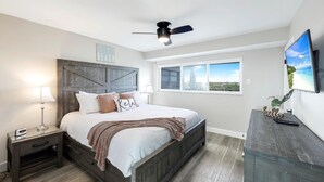 Un dormitorio con cama, mesita de noche, cómoda, ventilador de techo y ventana con vistas a la ciudad.