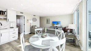 Una cocina moderna con armarios blancos, un comedor con mesa de vidrio y una sala de estar con sofá y televisor.