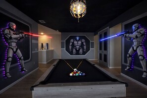A pool table with two Star Wars-themed decorations, a chandelier, and a wall with a large figure.