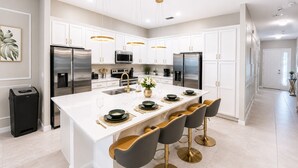 Una cucina moderna con mobili bianchi, elettrodomestici in acciaio inossidabile, un'isola centrale con sgabelli da bar e illuminazione a sospensione.