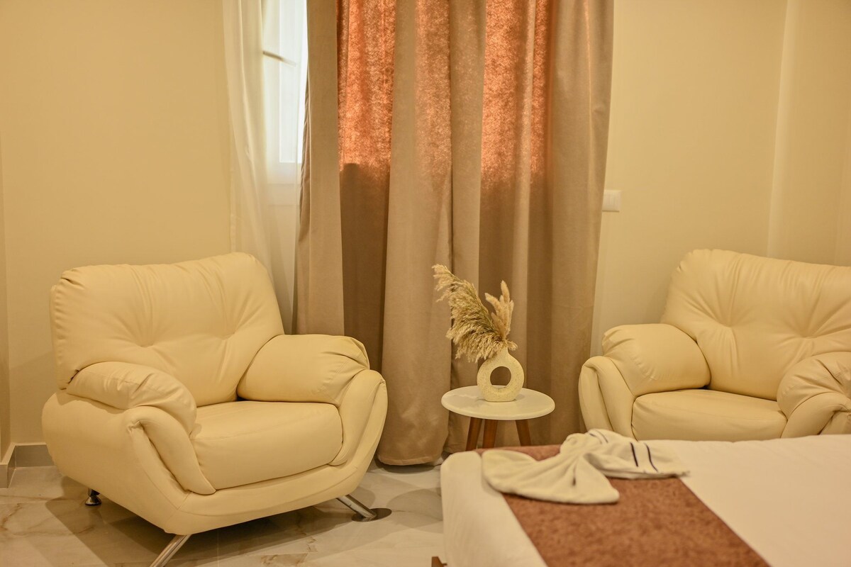 A hotel room with two cream-colored armchairs, a small round table with a decorative item, and a bed with a white comforter.