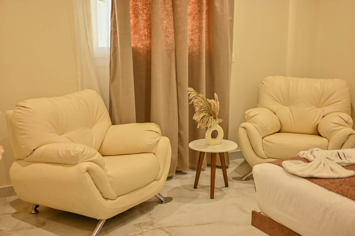 A room with two cream-colored armchairs, a small round table with a decorative item, and a bed with a white cover.