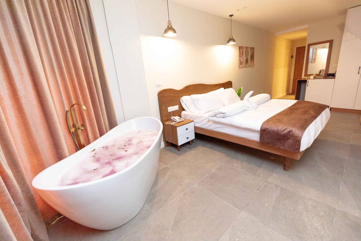 A hotel room with a large bathtub, a wooden bed, a nightstand, and a mirror.