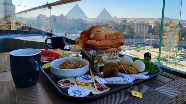 Un plateau de petit-déjeuner avec du pain, des œufs, du yaourt et du café, posé sur un balcon donnant sur une vue panoramique de la ville, avec des pyramides en arrière-plan.