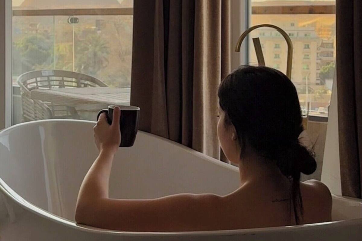 A person sitting in a bathtub with a view of a pyramid and cityscape through a window.