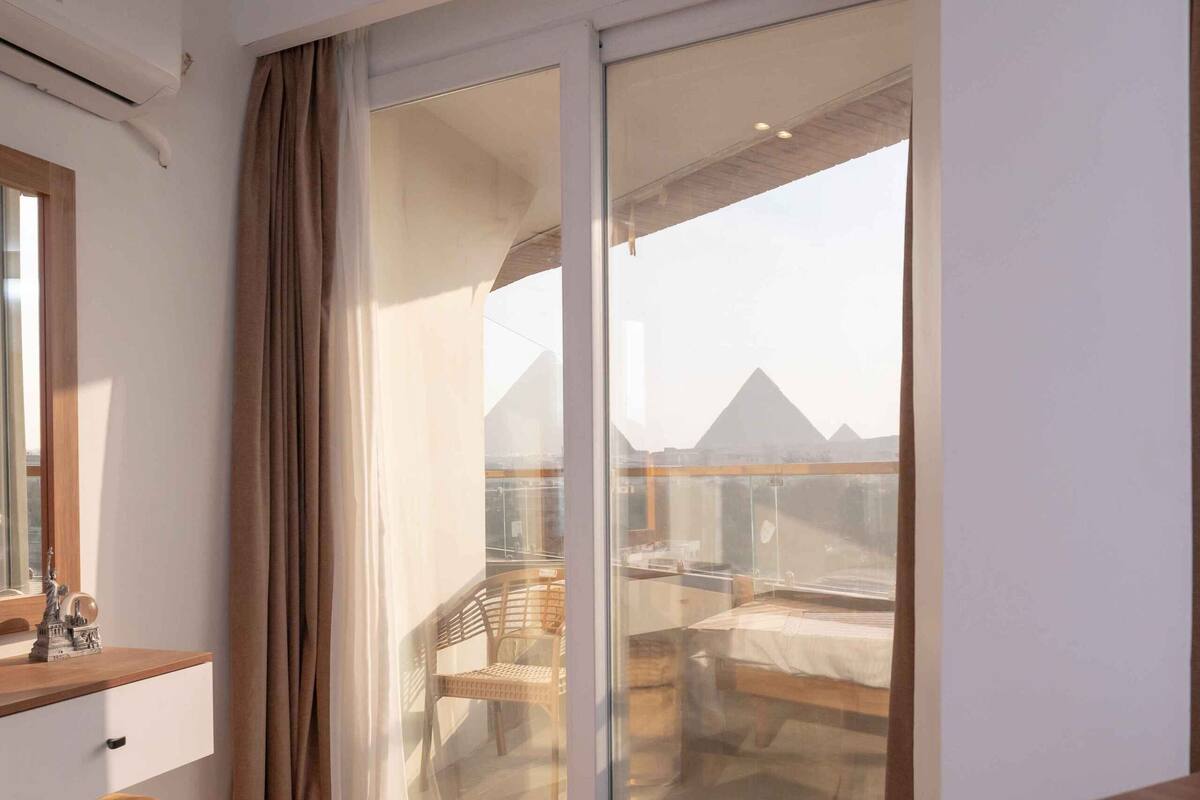 A hotel room with a bed, a dresser, a mirror, a window with curtains, and a view of pyramids.