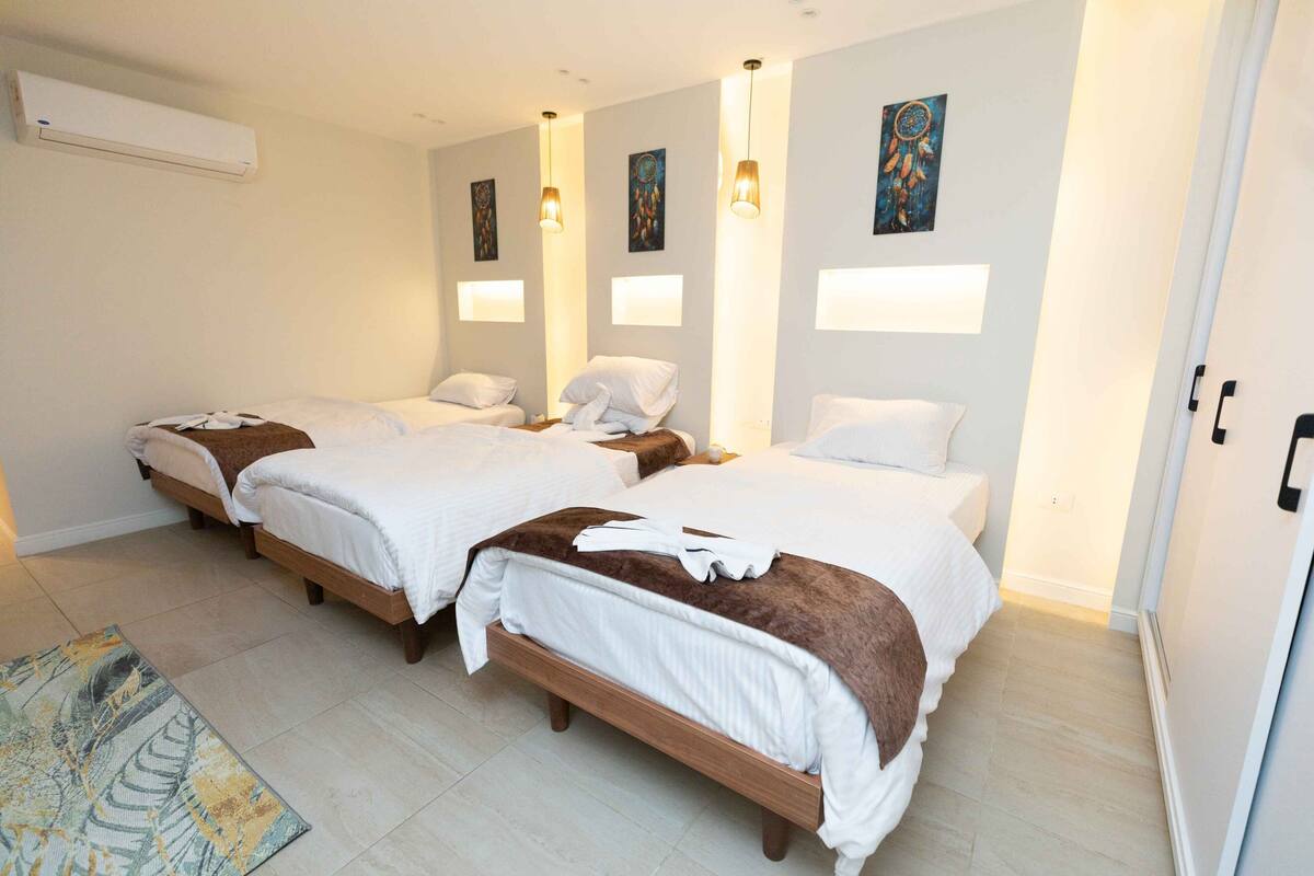 A hotel room with three beds, a large window, and a wall-mounted air conditioner.