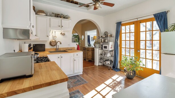 A kitchen with white cabinets, a wooden countertop, a ceiling fan, and a view through a large window with blue curtains.