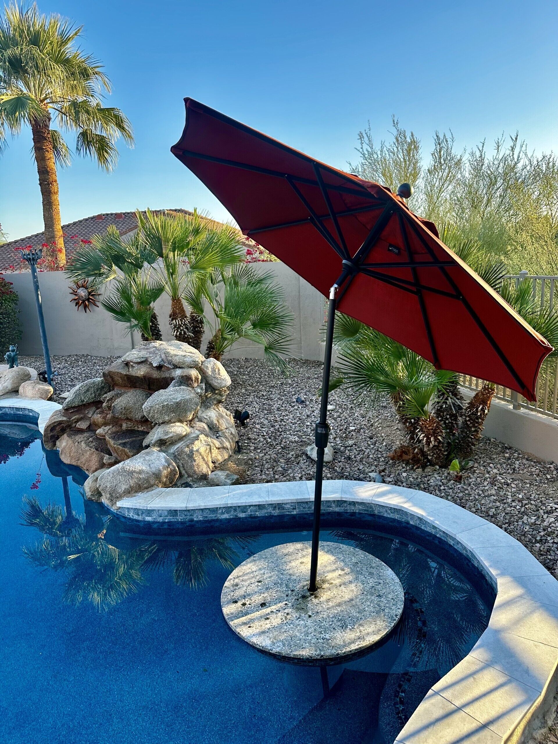 A swimming pool with a red umbrella, surrounded by rocks and palm trees.