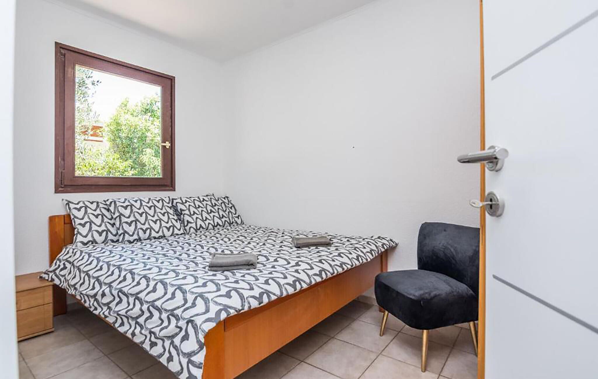 A bedroom with a bed, a chair, a nightstand, and a window with a view of greenery.