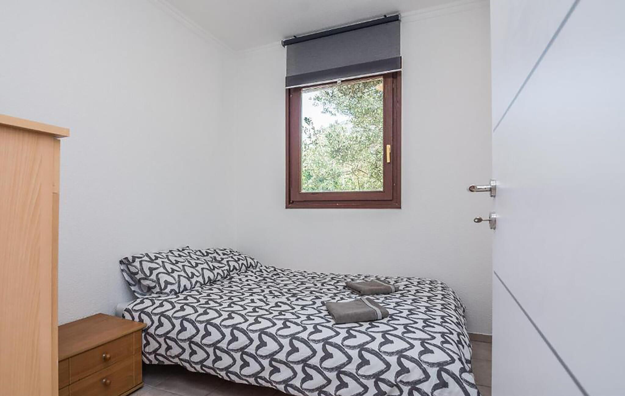 A bedroom with a bed, a wooden dresser, a window with a view of trees, and a door.