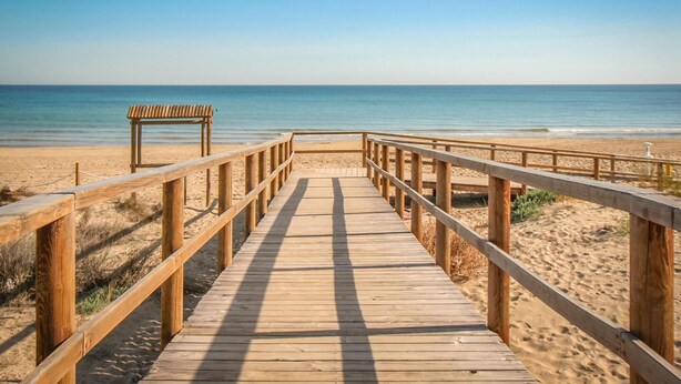 Un muelle de madera que conduce a una playa, con un banco y una barandilla.