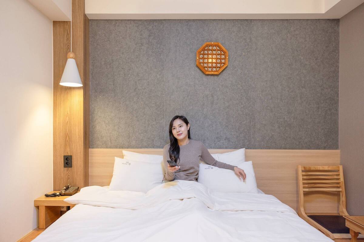 Une chambre d’hôtel avec une tête de lit en bois, un lit blanc, une table de chevet en bois avec une lampe et une chaise en bois.