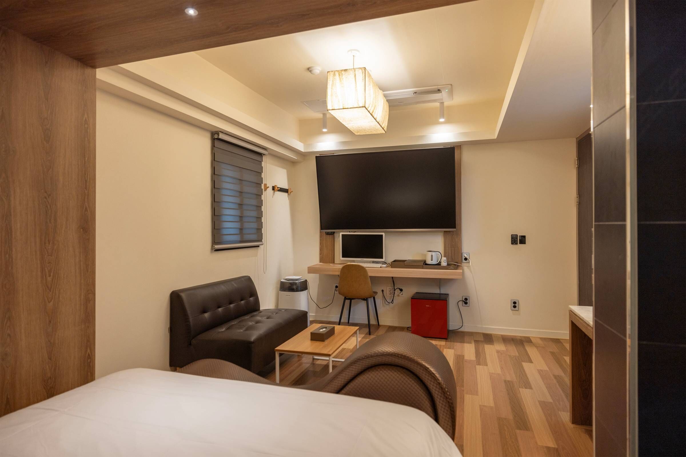 Una habitación de hotel compacta con una cama, un pequeño sofá, un escritorio con una computadora, un televisor y un mini refrigerador rojo.