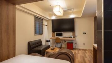 A compact hotel room with a bed, a small sofa, a desk with a computer, a TV, and a red mini-fridge.