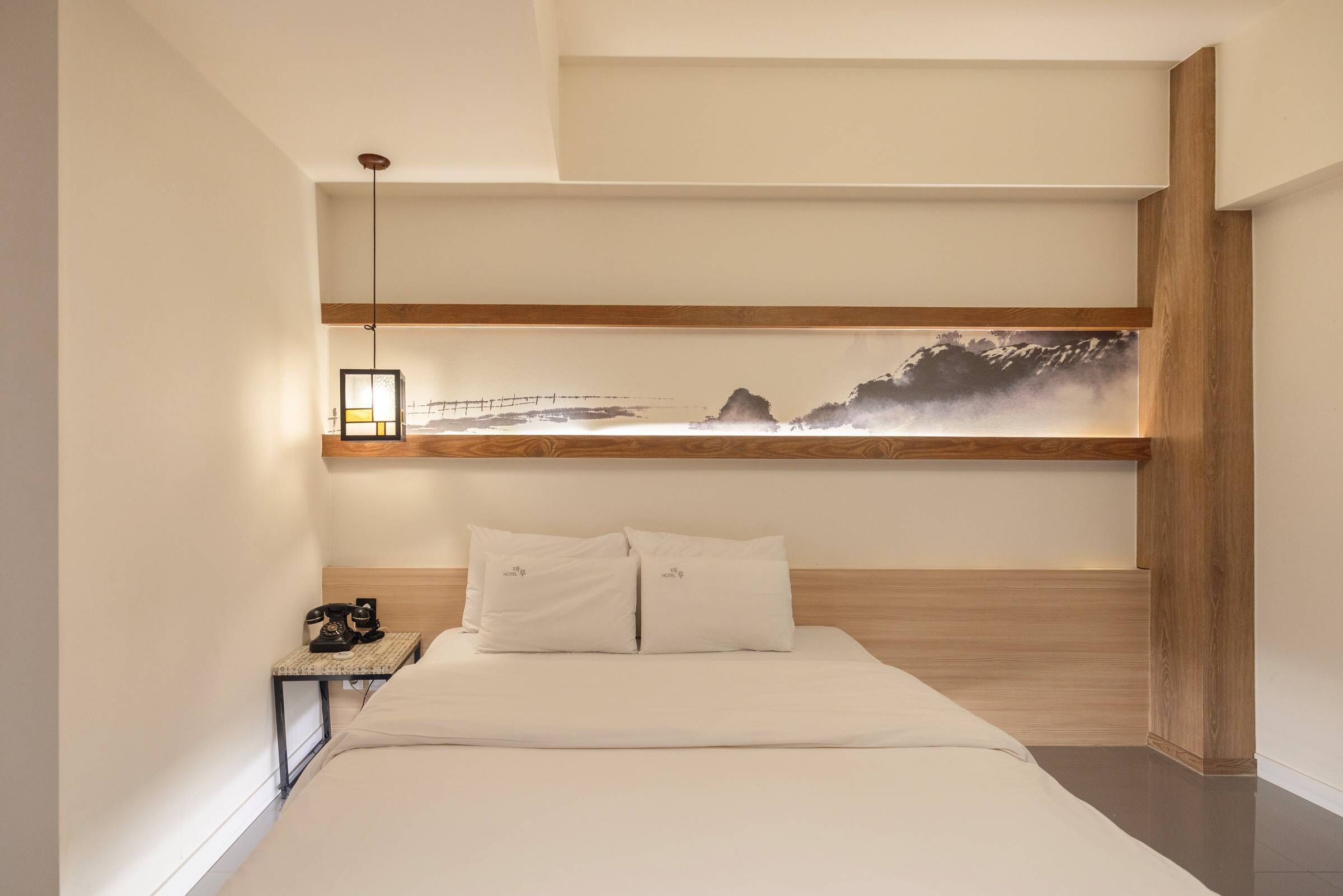 A modern bedroom with a wooden headboard, a bed with white linens, a nightstand with a lamp, and a wall-mounted shelf with a framed picture.