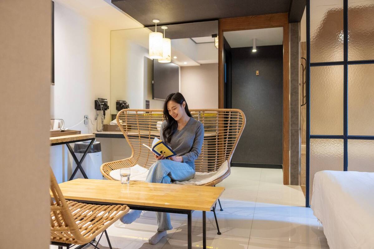 Une femme est assise sur une chaise en osier, lisant un livre dans une pièce moderne, où se trouvent une table en bois et un canapé blanc.