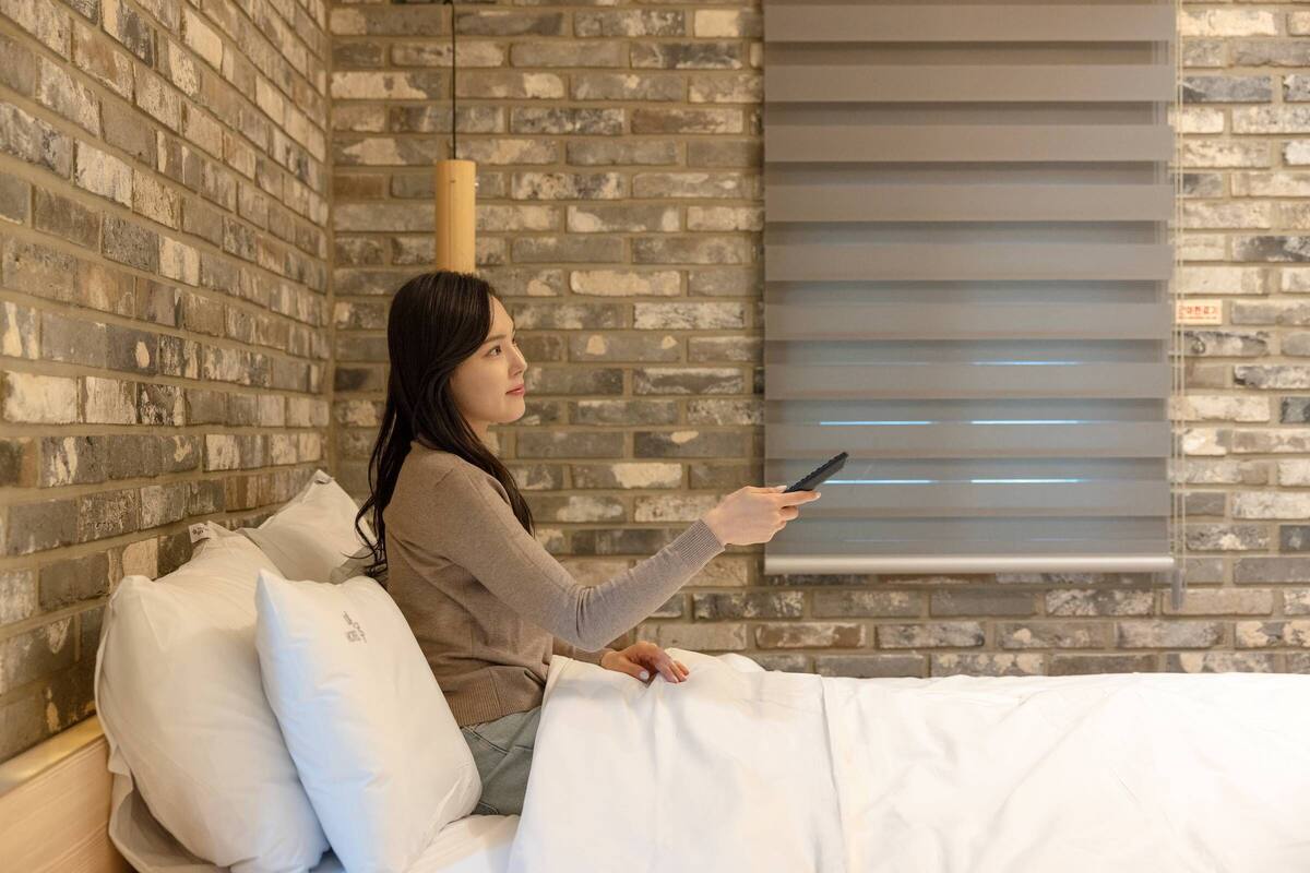 Une personne est assise sur un lit dans une pièce dont un mur est en briques et qui possède une fenêtre avec des stores.