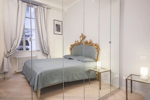A bedroom with a bed, a bedside table with a lamp, a window with curtains, and a radiator.