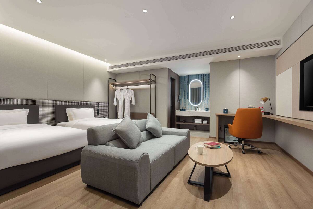 A modern hotel room with two beds, a grey sofa, a small round table, an orange office chair, a TV, a mirror, and a bathroom with a shower and toilet.