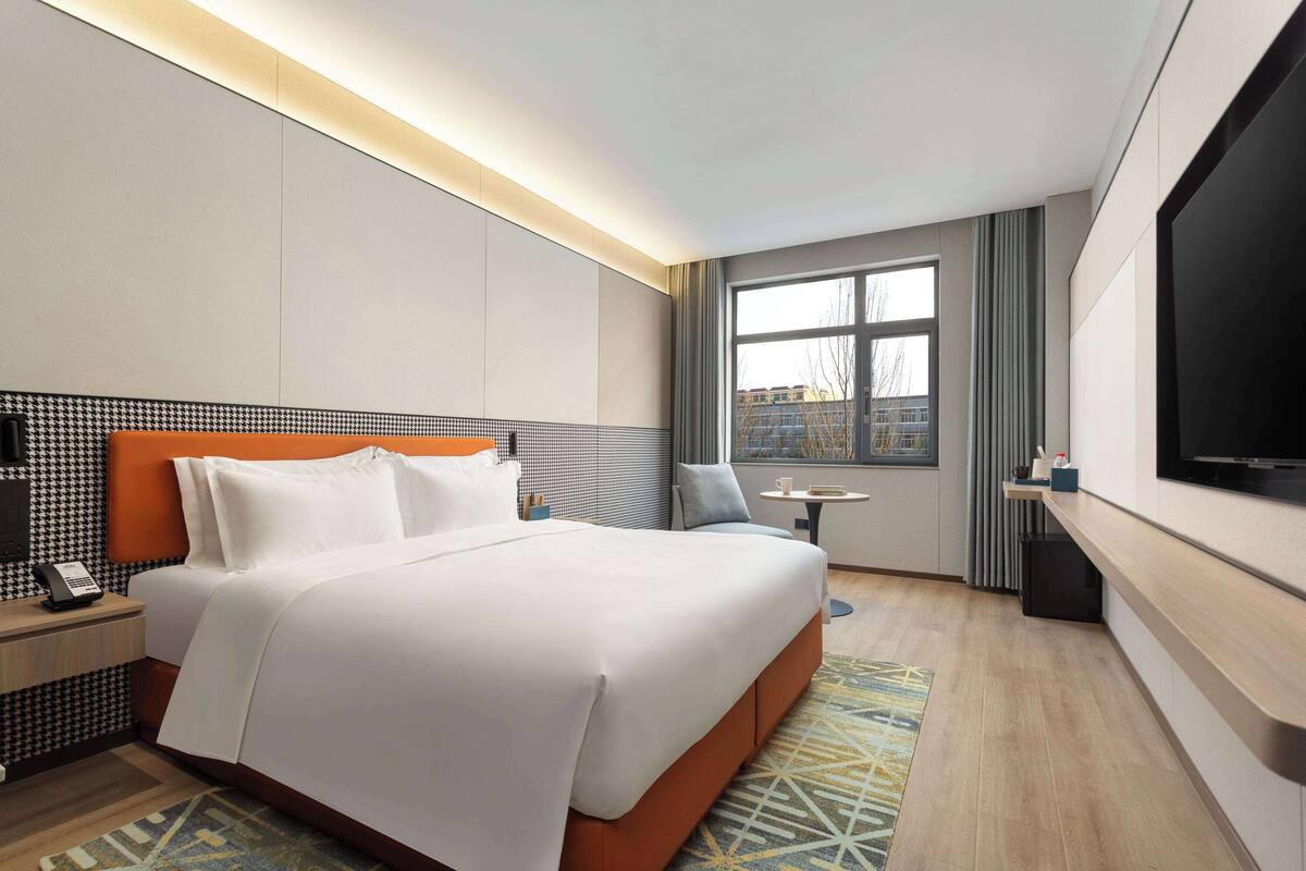 A hotel room with a large bed, a TV, a small table, and a window with a view of buildings.