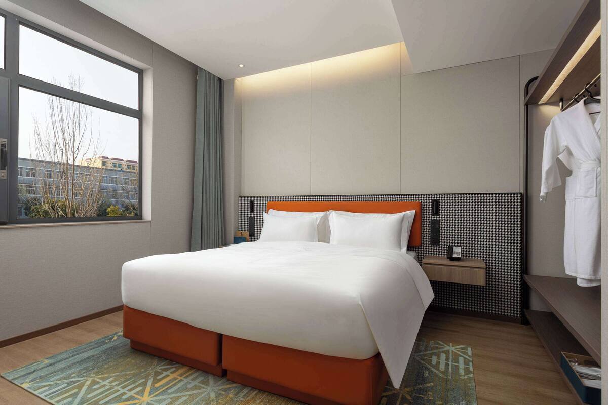 A modern hotel room with a large bed, a bedside table, a wardrobe, and a view of the city through a large window.