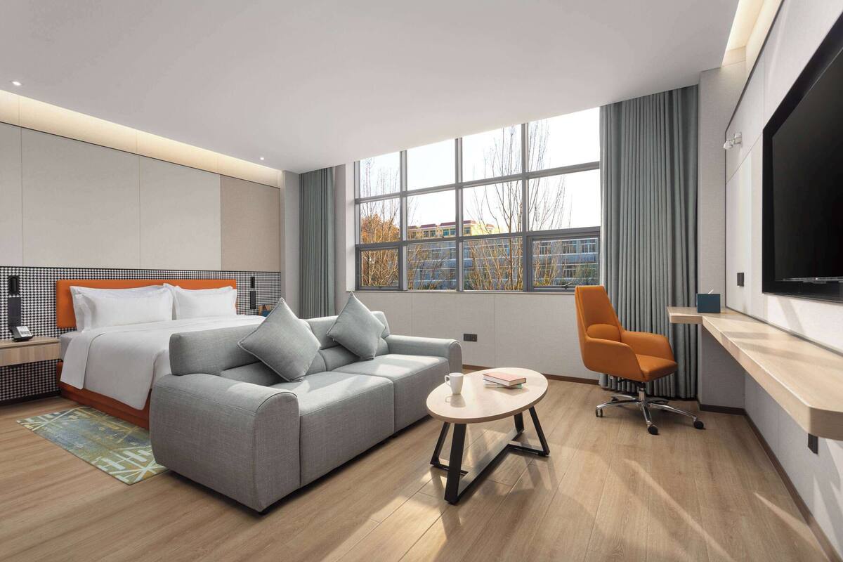 A modern hotel room with a bed, grey sofa, round coffee table, and a TV.