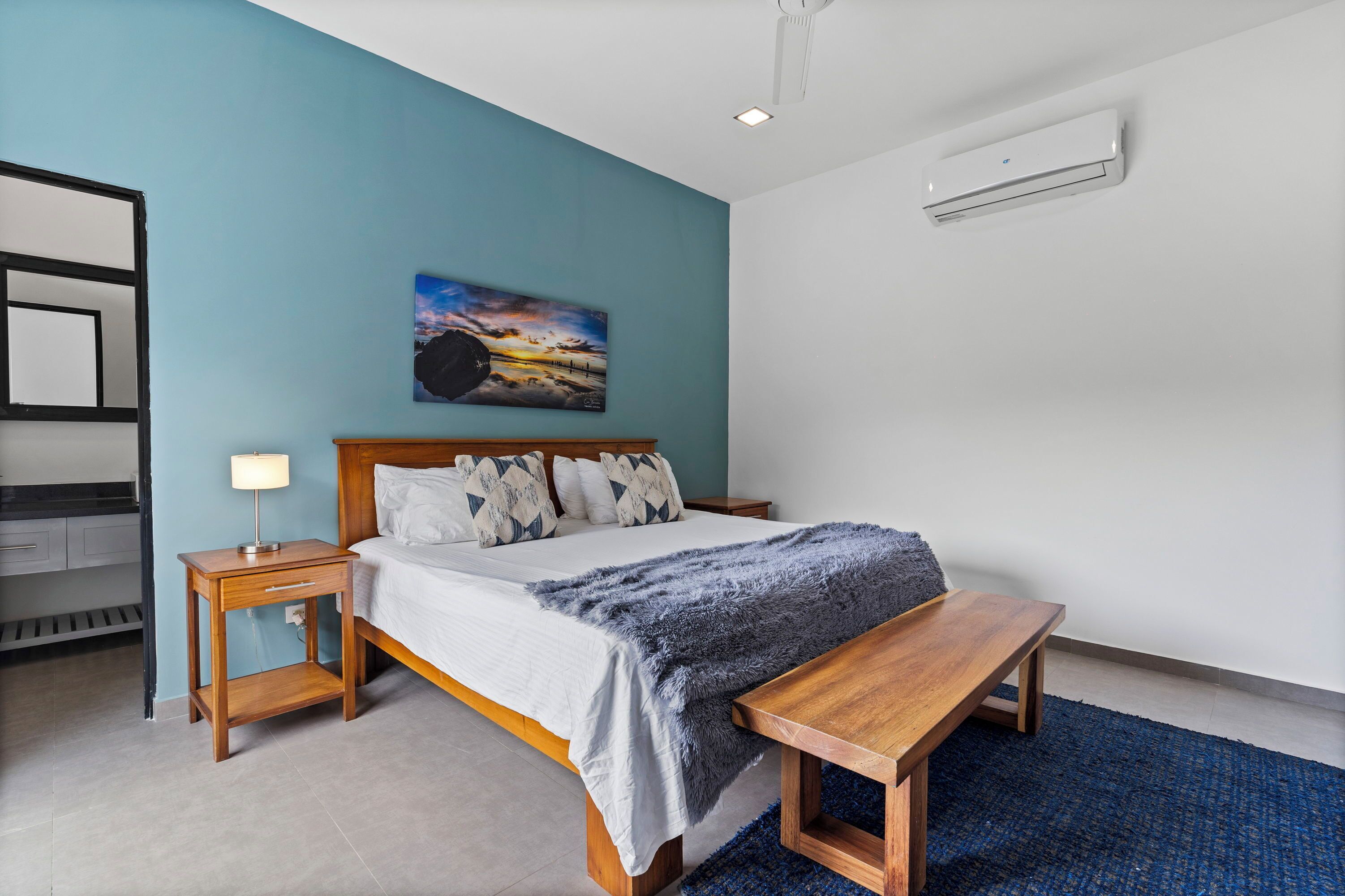 A bedroom with a bed, bedside tables, a bench, a painting, and an air conditioning unit.