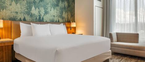A hotel room with a large bed, a headboard with a forest-themed wallpaper, a bedside table with a lamp, a sofa, and a large window with curtains.
