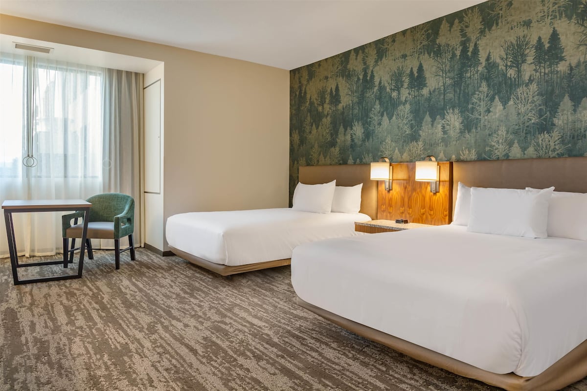 A hotel room with two beds, a desk, a chair, and a large window with curtains.