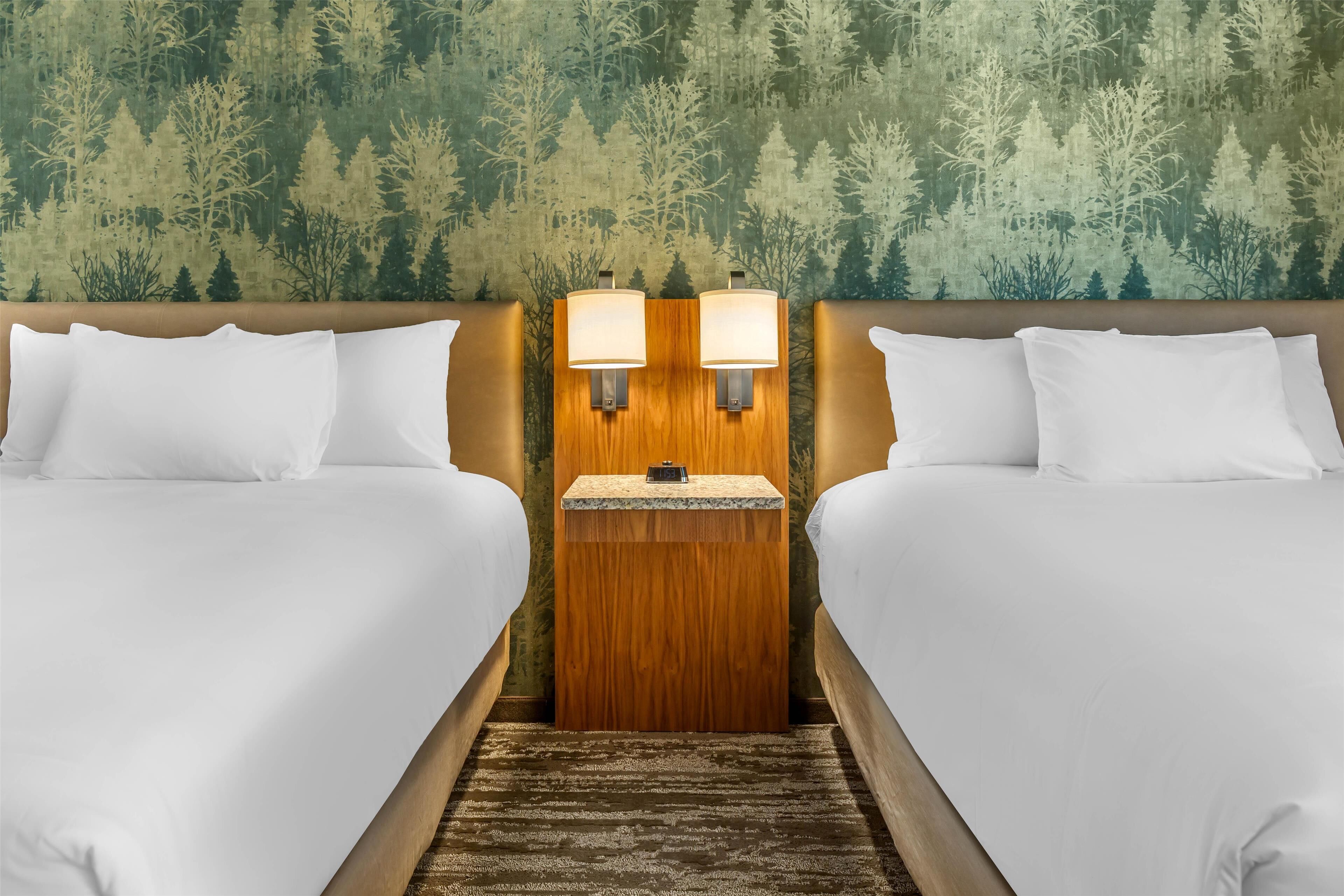 A hotel room with two beds, a wooden headboard, and a wall with a forest wallpaper.
