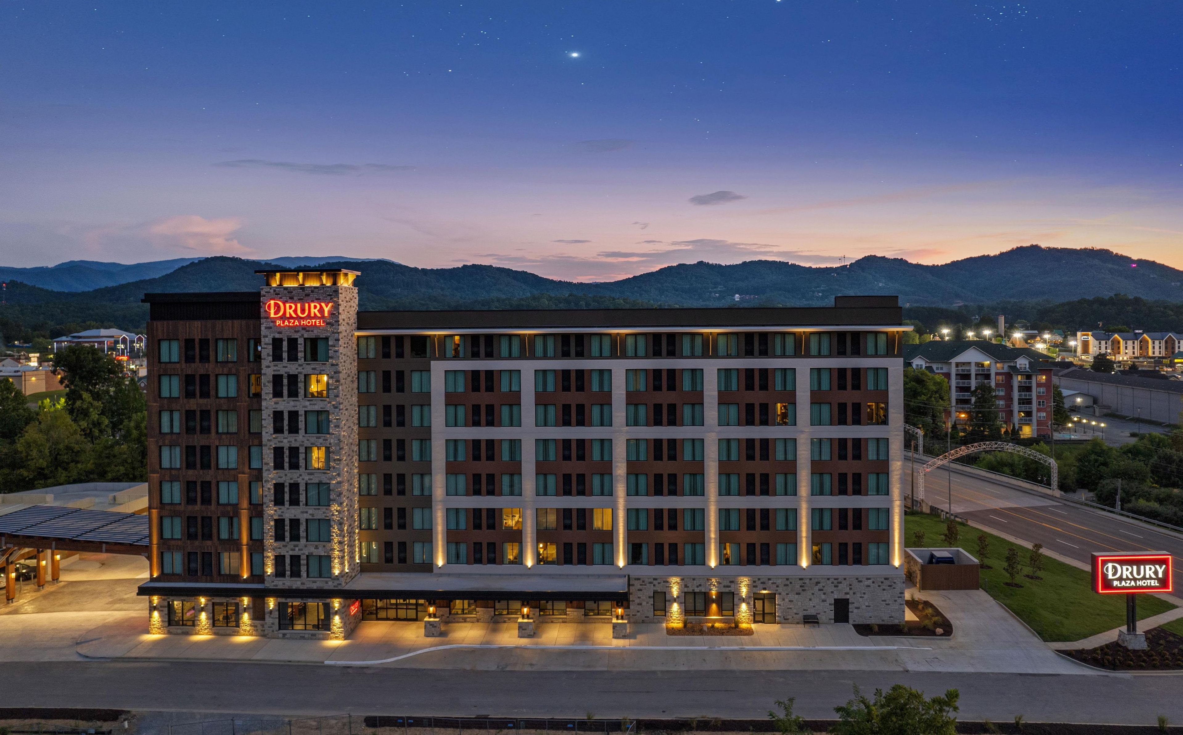 Un hôtel Drury Plaza à plusieurs étages, reconnaissable à son enseigne bien visible, entouré de montagnes et doté d’un parking bien éclairé.