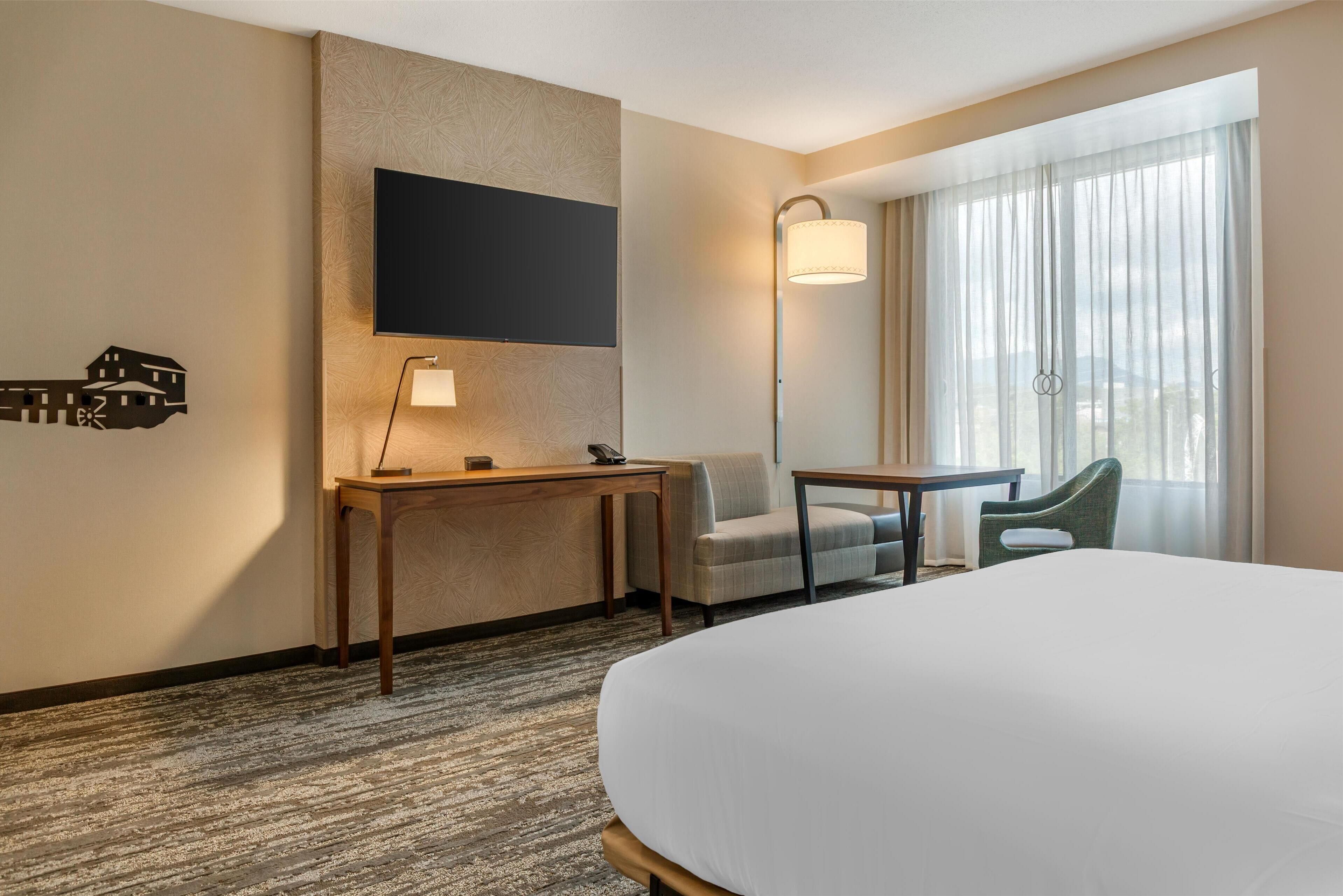 A hotel room with a large bed, a desk, a chair, a TV mounted on the wall, and a window with curtains.