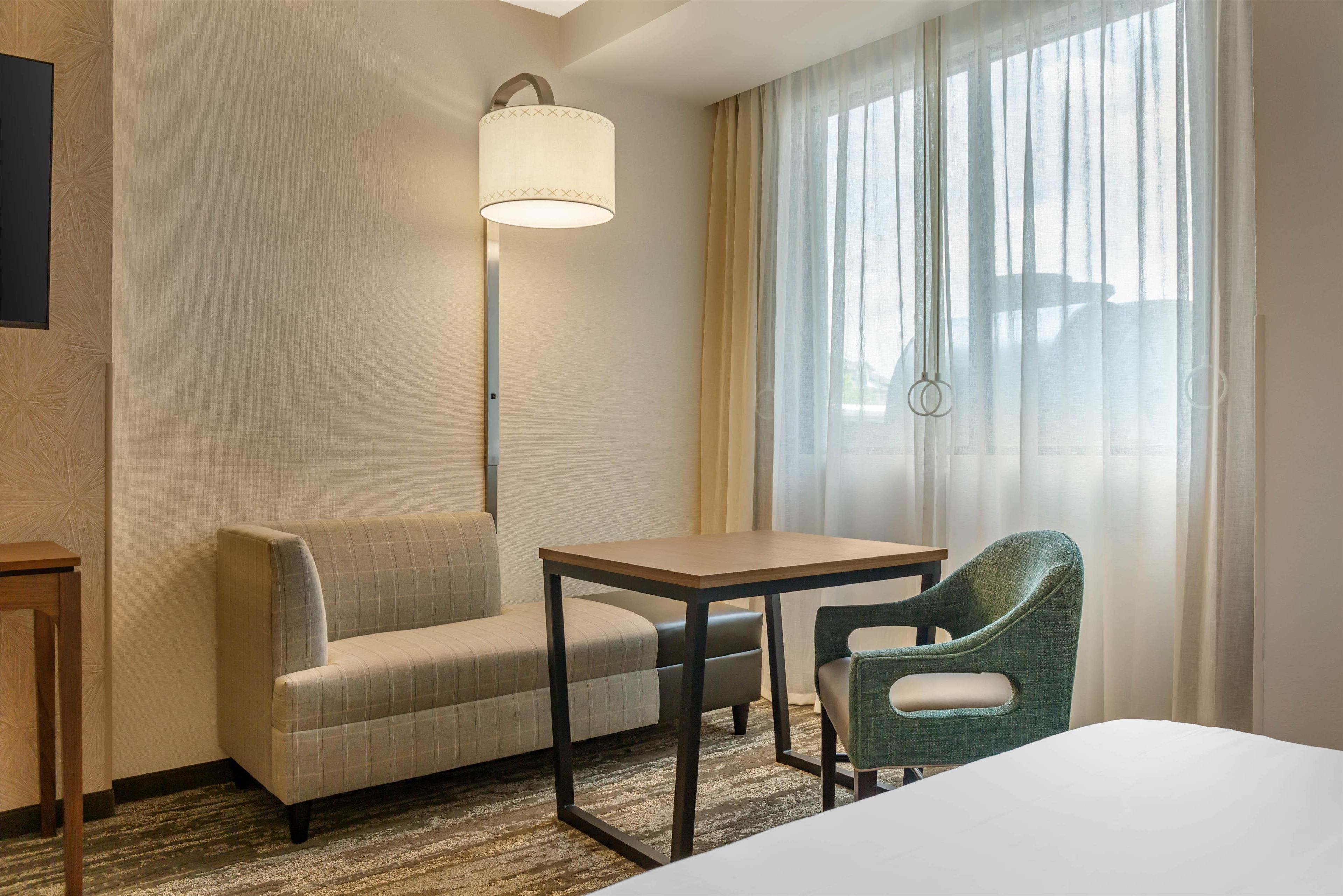 Habitación de hotel con sofá, una mesita, una silla, una lámpara y ventana con cortinas.