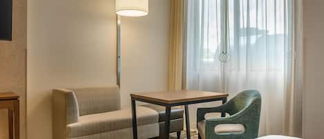 Een hotelkamer met een bank, een kleine tafel, een stoel, een lamp en een raam met gordijnen.