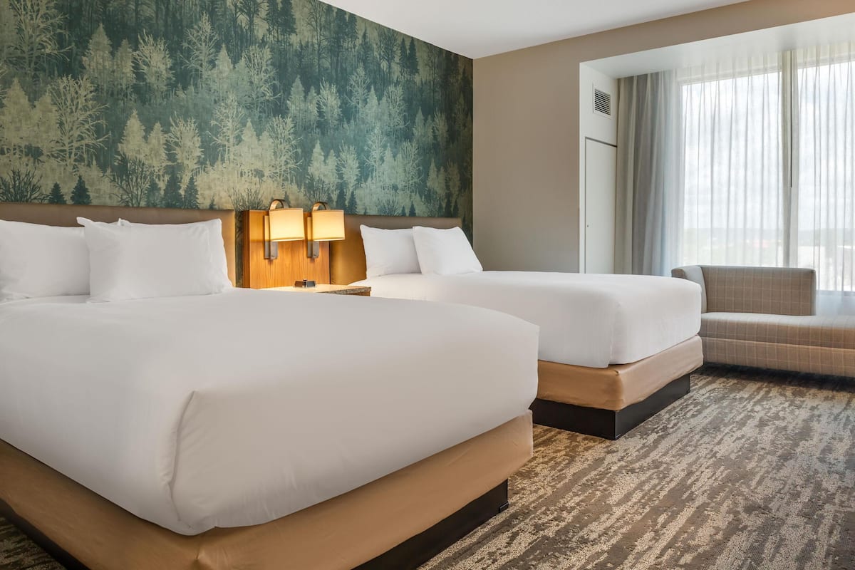 A hotel room with two beds, a sofa, a large window with curtains, and a forest-themed wallpaper.