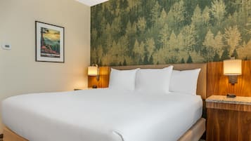 Una cama grande con sábanas blancas, dos mesitas de noche con lámparas, un cuadro en la pared y papel tapiz con motivos de bosque.