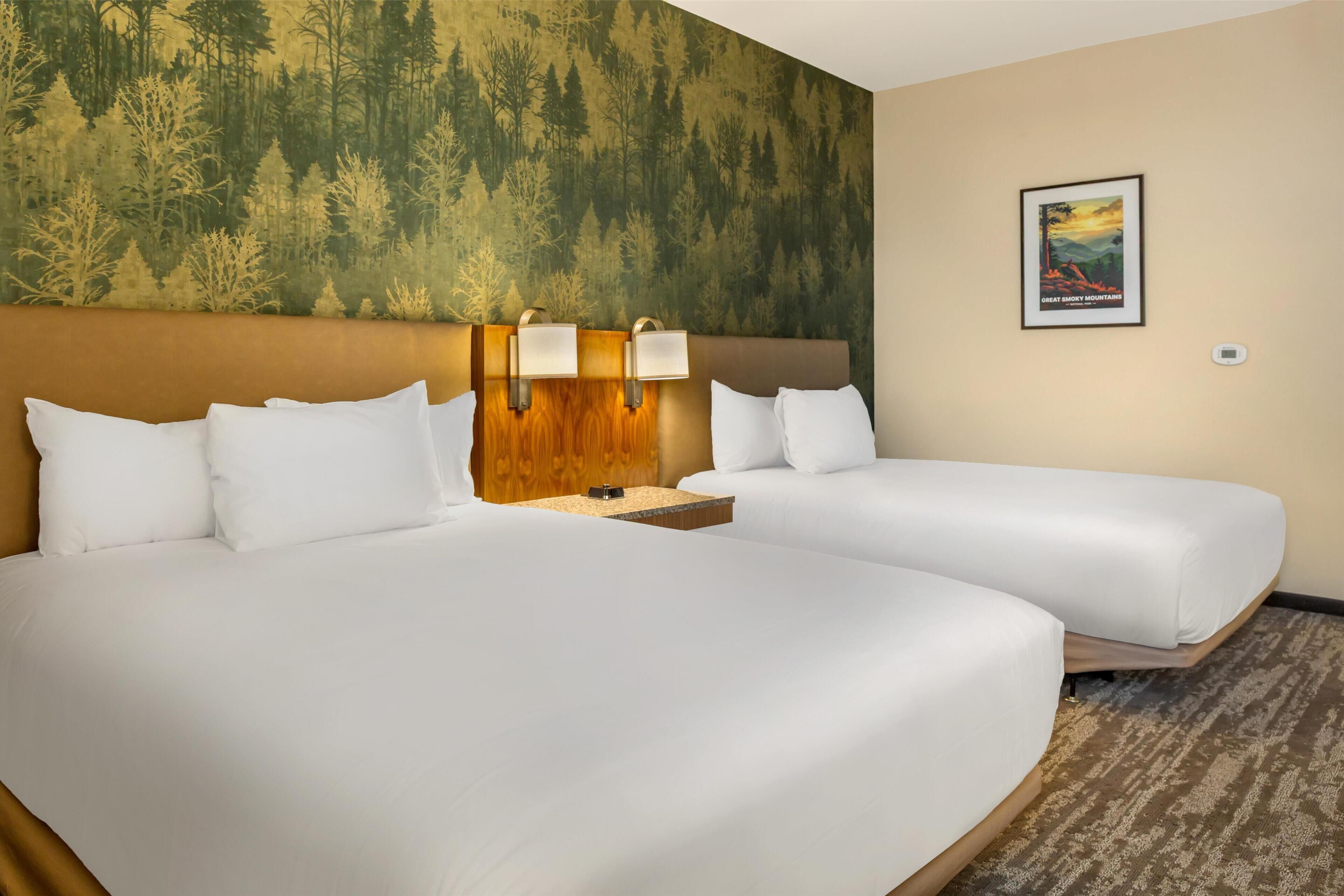 A hotel room with two beds, a headboard with wall-mounted lamps, a poster on the wall, and a carpeted floor.